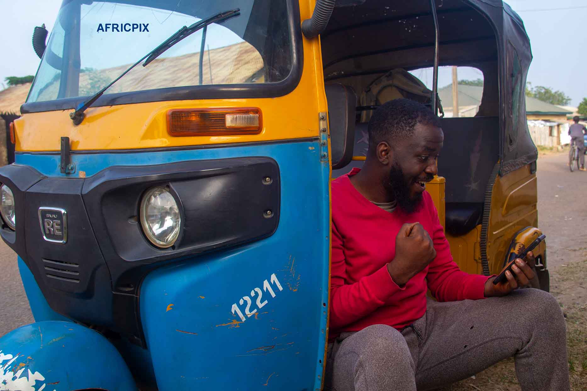 An excited African tricycle rider also known as Keke or Maruwa rider, holding his phone, smiling with joy after receiving good news.3